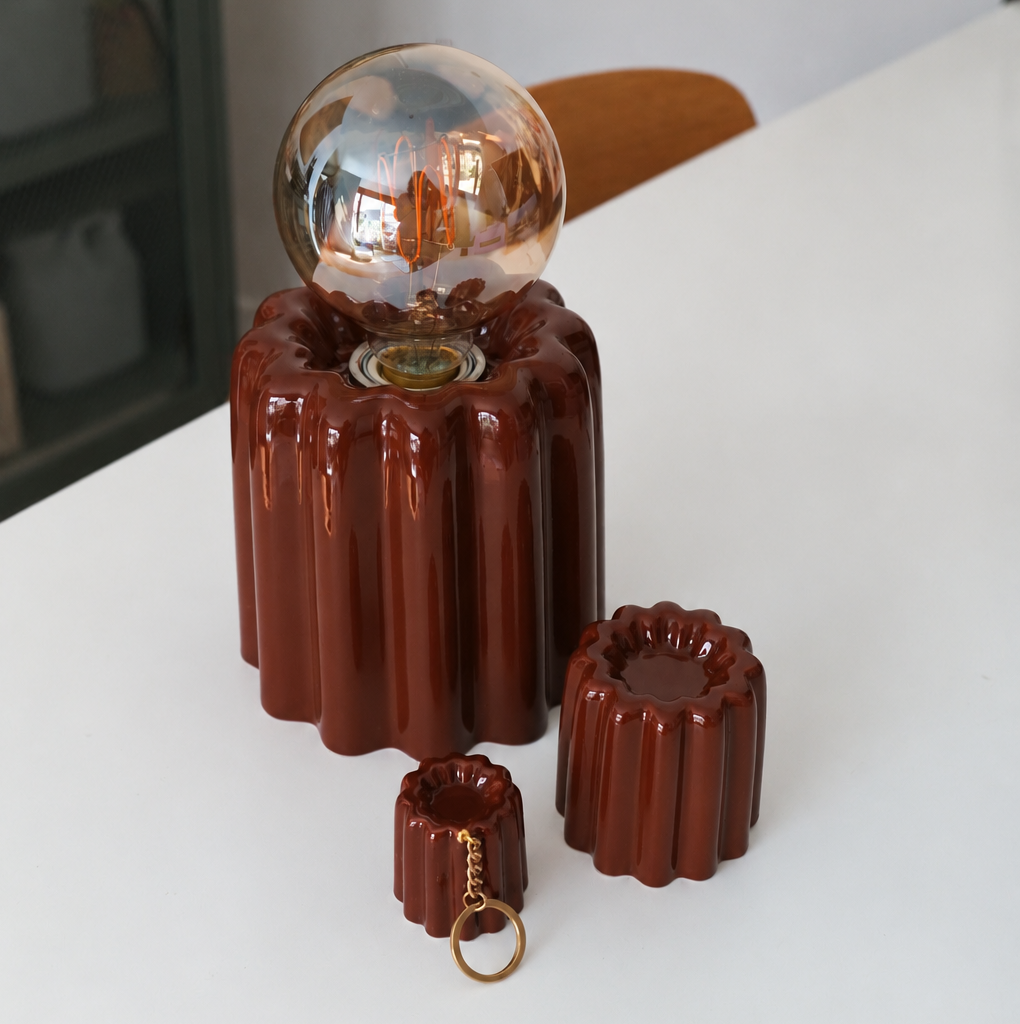 A glossy dark reddish-brown ceramic Canelé table lamp with a ribbed cylindrical base and a clear glass globe bulb, shown on a white tabletop with two matching small ceramic accessories in front.