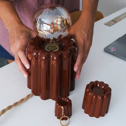 Sarah from BooBooclay holds a glossy dark brown canelé shape lamp with a clear bulb on a white table, with two matching brown canelés placed nearby.