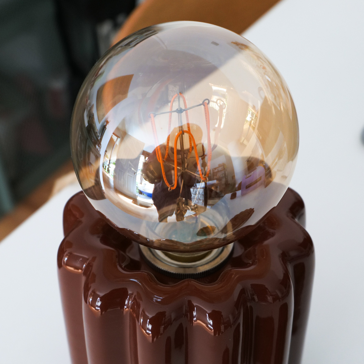 A close-up of a decorative table lamp with a large clear glass globe bulb and an orange filament, mounted on a glossy dark brown canelé shape ceramic.